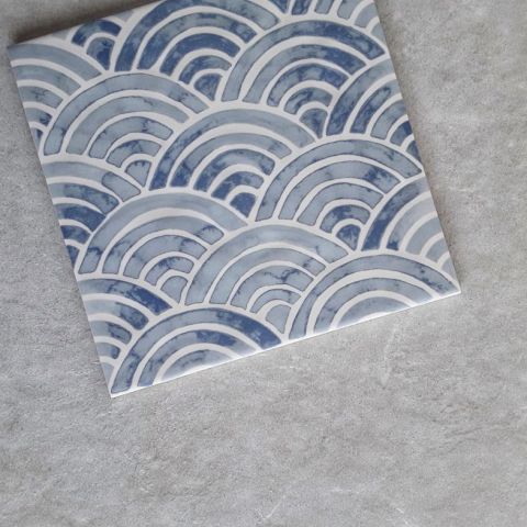 Handgefertigte Keramikfliese mit traditionellem Seigaiha Wellenmuster in Blautönen Dekorative Keramikfliese mit blau-weißem Wellenmuster im japanischen Stil auf grauem Untergrund